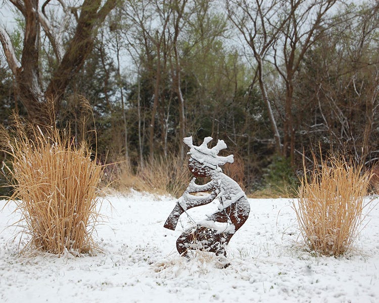 Snow falls on a Kokopelli sculpture and ornamental grasses in our customer Sarah's garden in Kansas.